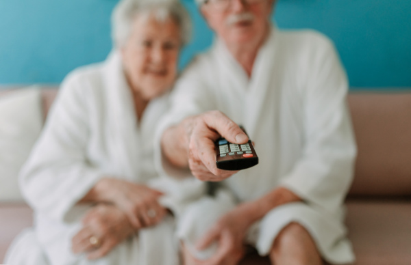 Happy senior couple sitting at a sofa in bathrobes and watching TV.