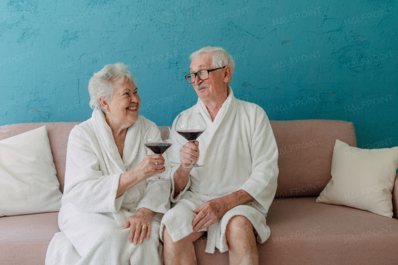 Happy senior couple sitting together in the bathrobe on sofa with glass of wine, having nice time at home.