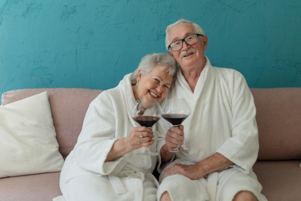 Happy senior couple sitting together in the bathrobe on sofa with glass of wine, having nice time at home.