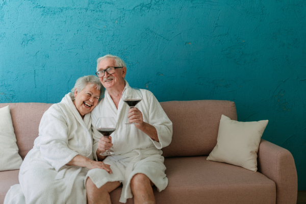 Happy senior couple sitting together in the bathrobe on sofa with glass of wine, having nice time at home.