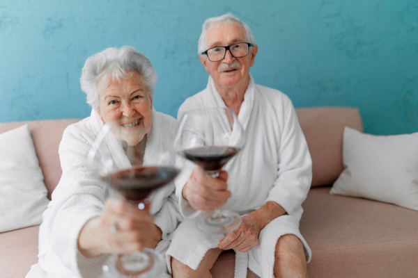 Happy senior couple sitting together in the bathrobe on sofa with glass of wine, having nice time at home.