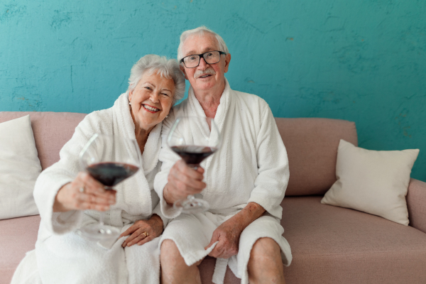 Happy senior couple sitting together in the bathrobe on sofa with glass of wine, having nice time at home.