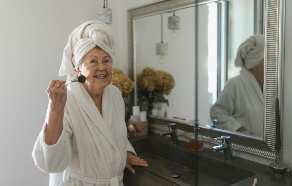 Senior woman standing in front of mirror in her bathroom and preparing her face with make up brush.