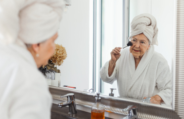 Senior woman standing in front of mirror in her bathroom and preparing her face with make up brush.