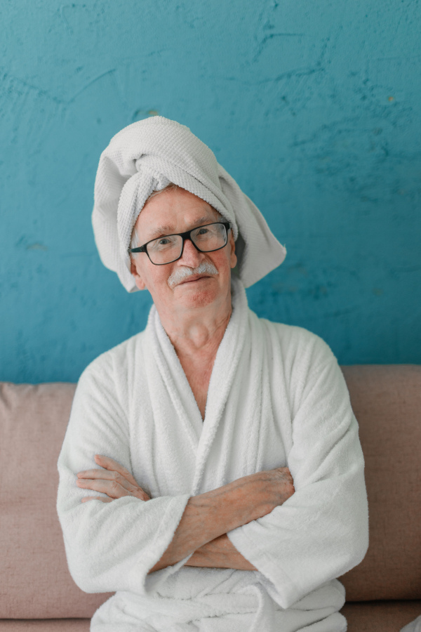 Happy senior man in glasses sitting at sofa in bathrobes and looking at camera.