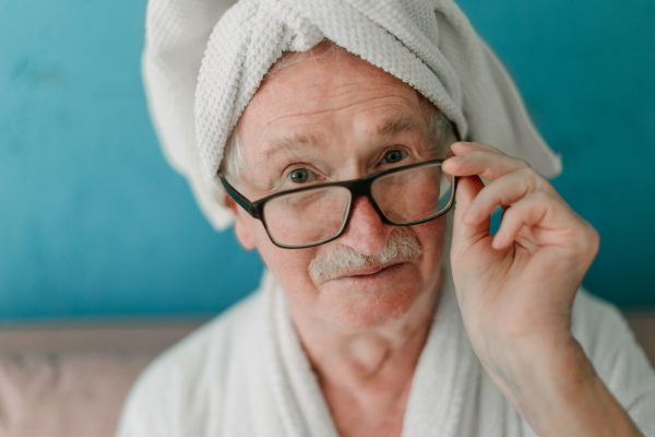 Happy senior man in glasses sitting at sofa in bathrobes and looking at camera.