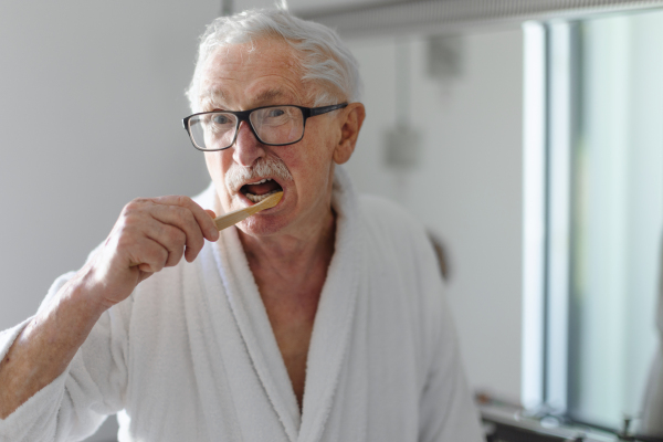 Senior man cleaning his teeth with a wooden brush in bathroom, sustainable lifestyle.