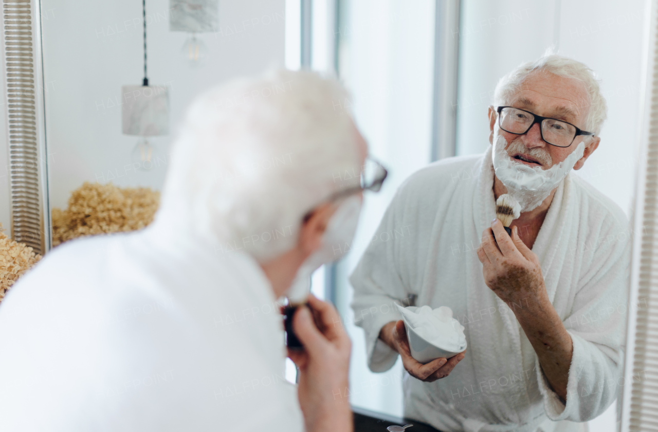 Senior man shaving his beard in bathroom, looking in the mirror. Morning routine concept.