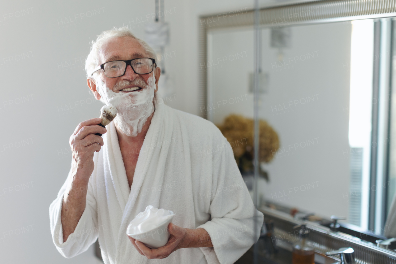 Senior man shaving his beard in bathroom, looking at camera. Morning routine concept.