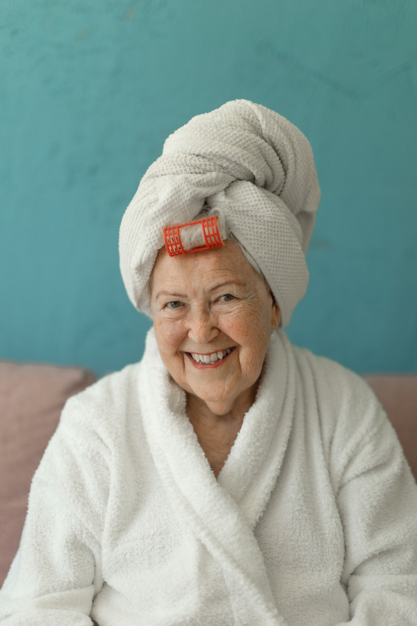 Happy senior woman with a plastic curler sitting at sofa in bathrobes and looking at camera.