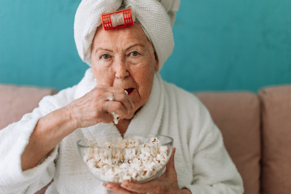 Happy senior couple sitting at a sofa in bathrobes and watching TV with popcorn.