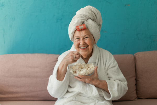 Happy senior woman sitting at a sofa in bathrobes and watching TV with popcorn.