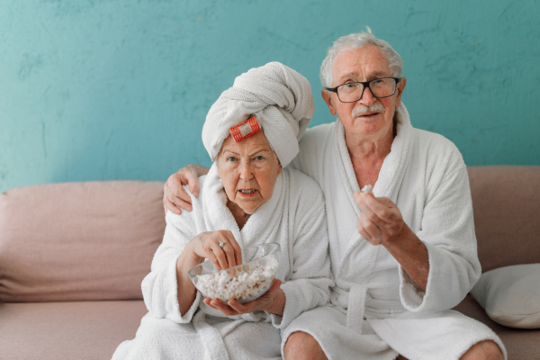 Happy senior couple sitting at a sofa in bathrobes and watching TV with popcorn.