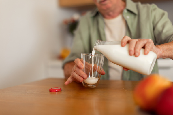 Close up of senior man sitting in kitchen and pouring milk in a glass. Healthy lifestyle concept.