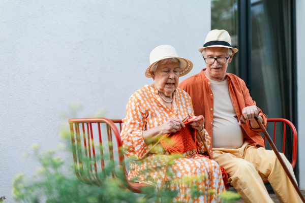 Senior woman sitting in the garden on the bench with his husband and knitting scarf.