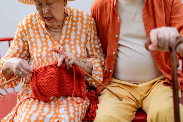 Senior woman sitting in the garden on the bench with his husband and knitting scarf.