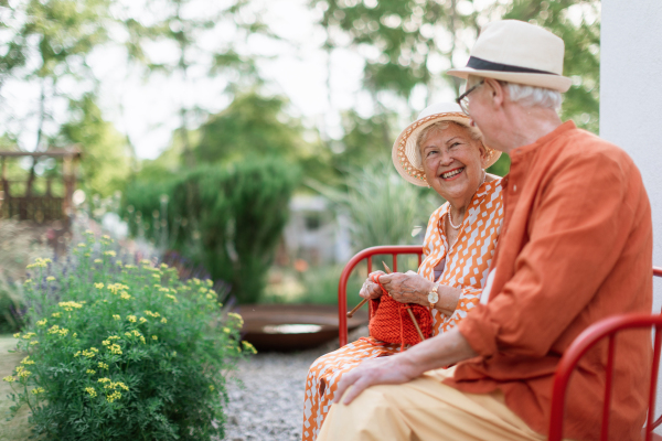 Senior woman sitting in the garden on the bench with her husband and knitting scarf.