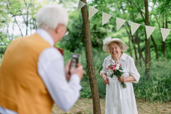 Senior couple having marriage in nature during a summer day, senior man taking photo of his wife.