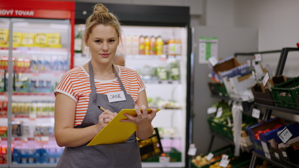 A young shop assistant in supermarket in vegetable shell, in bakcground is her colleague filling stock.