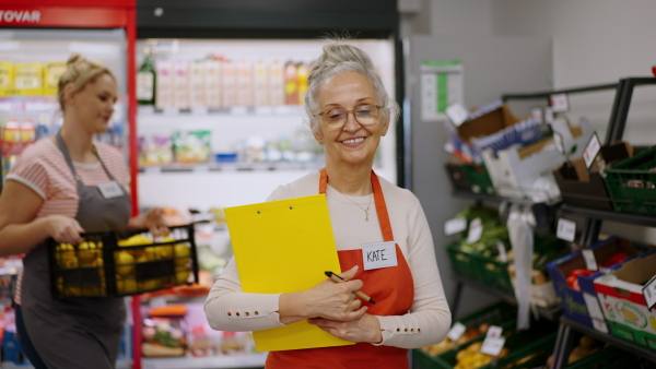 A confident senior shop assistant in supermarket in vegetable shell, in bakcground is her colleague filling stock.