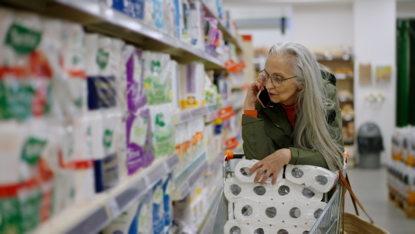 A senior woman buying toliet paper in supermarketm and calling on cellphone.