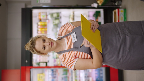 A young shop assistant in supermarket in vegetable shell, in bakcground is her colleague filling stock.