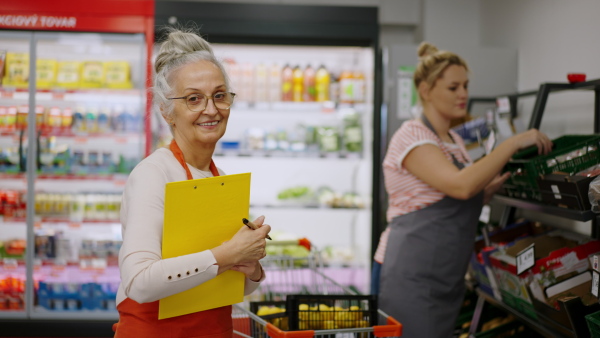 A confident senior shop assistant in supermarket in vegetable shell, in bakcground is her colleague filling stock.