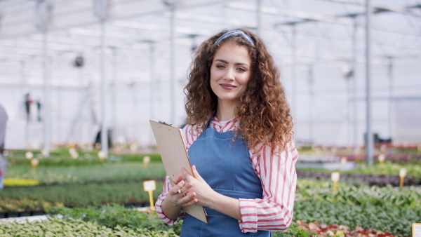 Young woman with clipboard working in garden centre, looking at camera.