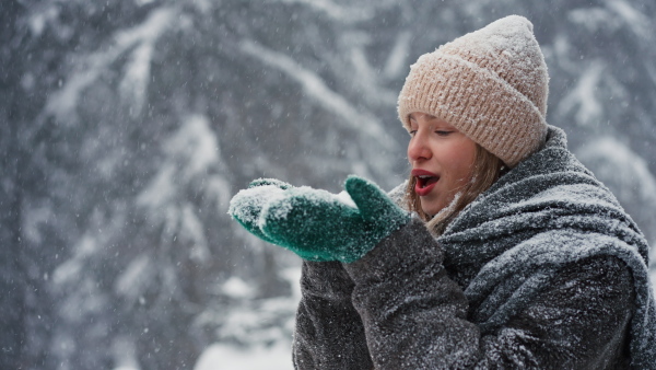 A happy young woman blowing snow from hands in winter under falling snow in nature