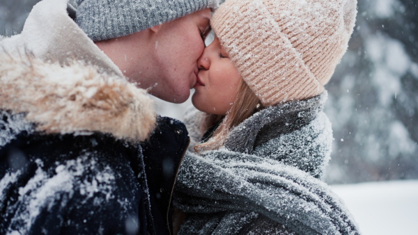 A close-up of young couple hugging and kissing in winter under falling snow in nature.