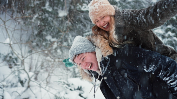 Happy young couple in love walking in wintery nature under falling snow, young man giving piggyback to his girlfriend in a snowy forest.