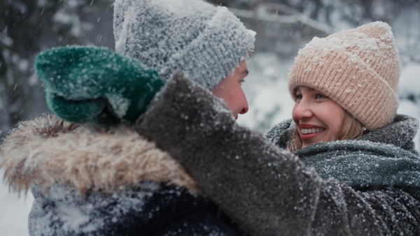 A close-up of young couple hugging and kissing, smiling, whispering something in winter under falling snow in nature.