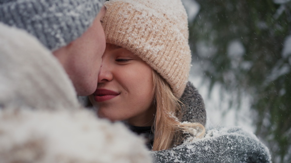 A close-up of young couple hugging and kissing, smiling, whispering something in winter under falling snow in nature.