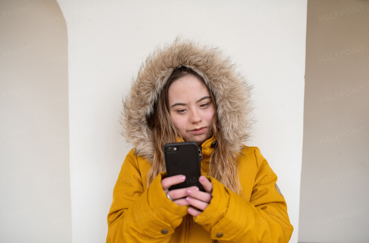 A happy young woman with Down syndrome weraing parka, leaning the wall and using smartphone.