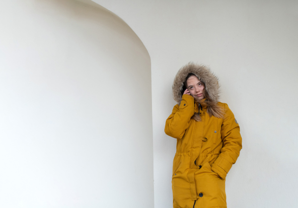 A thoughtful young woman with Down syndrome weraing parka, leaning the wall and looking at camera.