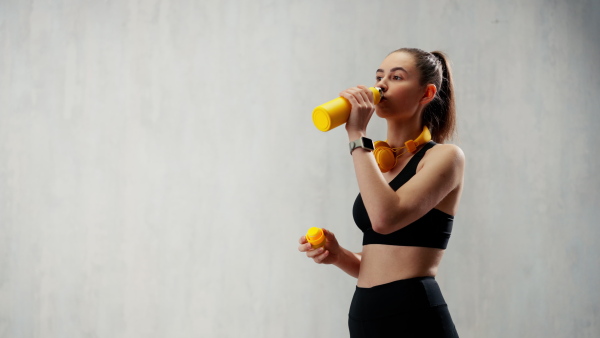 A young sporty woman in sportswear with wireless headset drinking from bottle over grey background