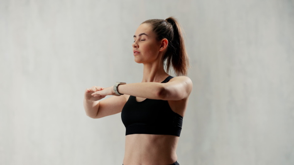 A young sporty woman in sports clothes stretching arms against grey background, copy space