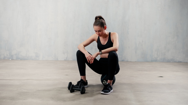 A tired young sporty woman having rest after workout, sitting on floor at gym , over grey background.