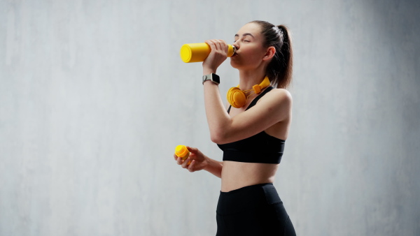 A young sporty woman in sportswear with wireless headset drinking from bottle over grey background