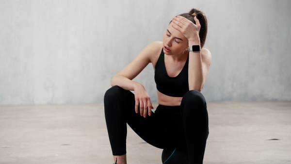 A tired young sporty woman having rest after workout, sitting on floor at gym , over grey background.