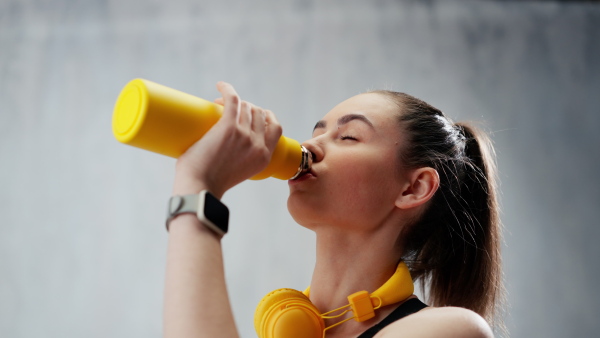 A young sporty woman in sportswear with wireless headset drinking from bottle over grey background