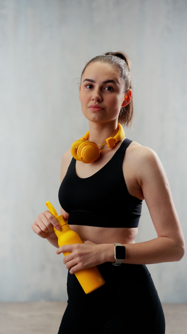 A young sporty woman in sportswear with wireless headset and bottle over green background looking at camera.