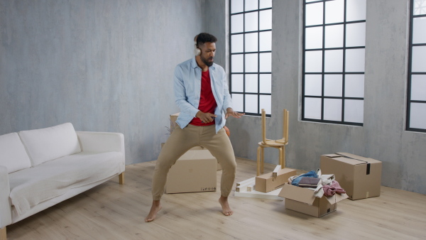 A happy young man with headphones dancing and celebrating moving home surrounded by packing boxes, new living concept.