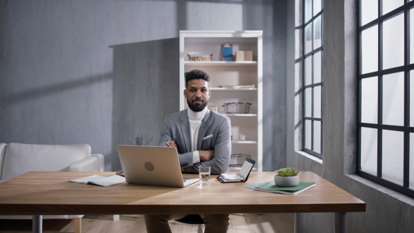 A happy young african american businessman working on a laptop indoors in office, looking at camera.