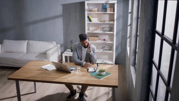 A happy young businessman using laptop and looking in diary while talking on mobile phone indoors in office