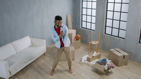 A happy young man with headphones dancing and celebrating moving home surrounded by packing boxes, new living concept.