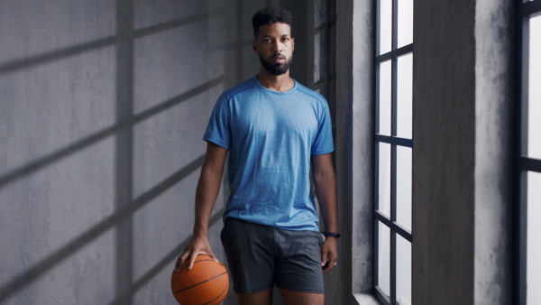 A young African American basketball player standing indoors at gym, looking at camera.