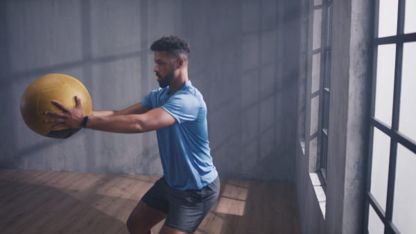 A young African American sportsman standing and lifting a medicine ball indoors, workout training concept.