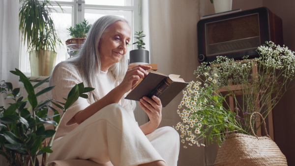 A relaxed senior woman sitting on floor and reading book indoors at home.