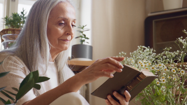 A relaxed senior woman sitting on floor and reading book indoors at home.